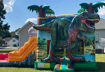 Bring excitement to your next event with the Dinosaur-Bounce-House-and-Slide-Rental, featuring a fun dinosaur design and a yellow slide on the left—set up outdoors on vibrant grass near houses under a blue sky. Reserve your Dinosaur Bounce House and Slide combo rental in Wilmington, NC today from Funtime Inflatables!. Bounce house rentals Wilmington NC - Funtime Inflatables NC