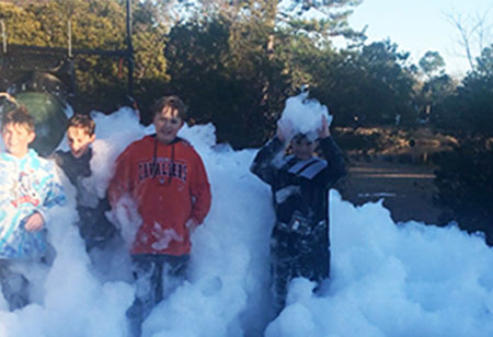 Four children stand outdoors surrounded by thick white foam at a lively Wilmington, NC foam party, with the Dinosaur Bounce House and Slide Rental visible nearby, plus trees and blue sky in the background. Bounce house rentals Wilmington NC - Funtime Inflatables NC