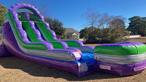 The Dual Lane Purple Beach inflatable slide is set up on a grassy yard with trees and houses in the background under a clear blue sky. Bounce house rentals Wilmington NC - Funtime Inflatables NC