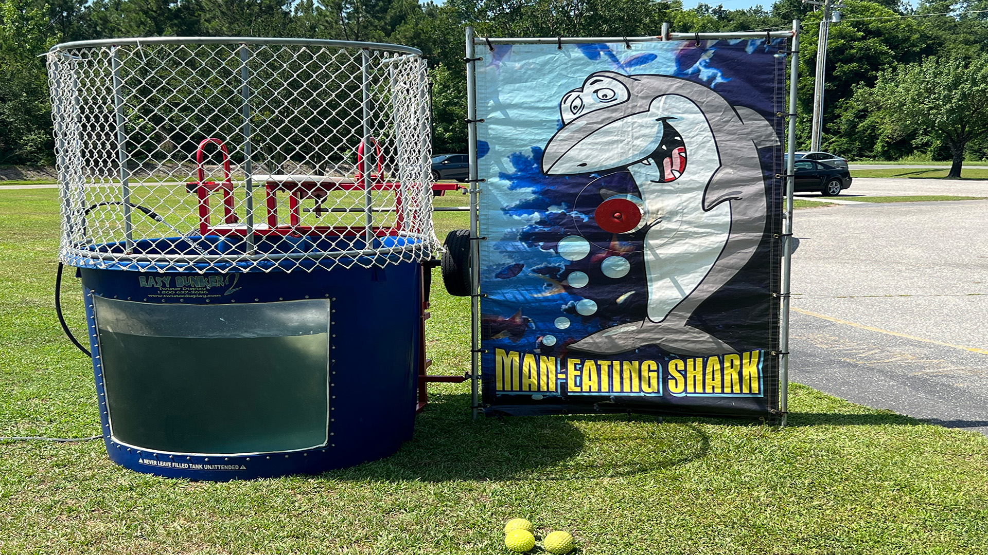 A cheerful Dunk Booth 1 rental in Wilmington, NC includes a dunk tank with a cage, water, and a target situated beside a sign featuring a cartoon shark and the phrase “Man-Eating Shark,” all arranged on grass with several yellow balls scattered nearby. Bounce house rentals Wilmington NC - Funtime Inflatables NC