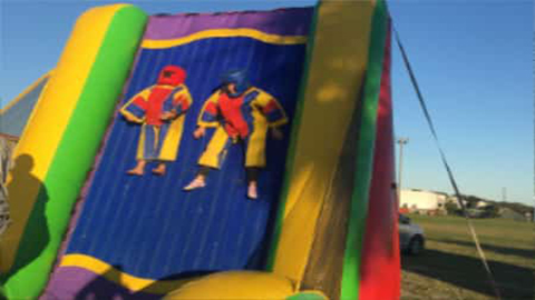 Two people in Velcro suits are stuck to a vertical Inflatable Retro Velcro Wall at an outdoor event, with grass and buildings visible in the background—perfect for anyone searching for a fun funtime-inflatables-NC-velcro-wall-rental experience in Wilmington, NC. Bounce house rentals Wilmington NC - Funtime Inflatables NC