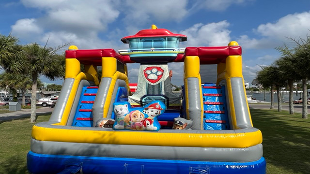 A vibrant, Paw Patrol-themed inflatable obstacle course featuring playful cartoon character figures at the front, set up on lush grass with picturesque palm trees and a marina in the background beneath a partly cloudy sky—perfect for bringing adventurous fun to your next outdoor event. Bounce house rentals Wilmington NC - Funtime Inflatables NC
