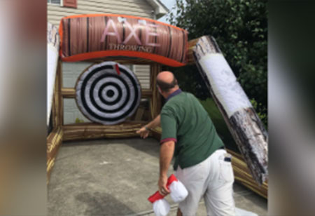 A man in a green shirt throws a toy axe at a target in the Funtime-Inflatables-NC-Axe-Throwing-rental inflatable game, set up outdoors on a driveway in Wilmington, NC. Bounce house rentals Wilmington NC - Funtime Inflatables NC