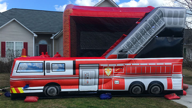 The fireTruck-funtime inflatable rentals bounce house, designed in the shape of a red fire truck, is set up on grass outside a house beneath a partly cloudy sky. Bounce house rentals Wilmington NC - Funtime Inflatables NC