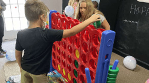 Two children play a large vertical Connect Four game indoors, with balloons and a chalkboard in the background—perfect inspiration if you’re considering the funtime Inflatables Rentals Giant Connect Four Rental in Wilmington, NC. Bounce house rentals Wilmington NC - Funtime Inflatables NC